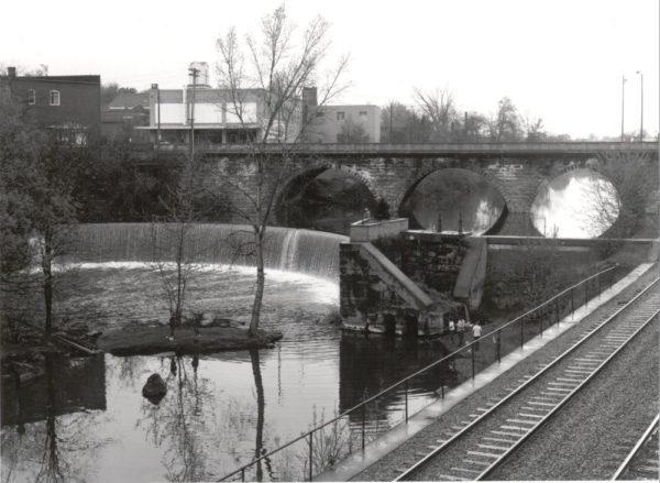 black and white photo of dam with water spilling over, wall and railroad to river left