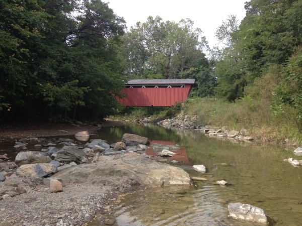 rocks and water in foreground, red covered bridge in background.