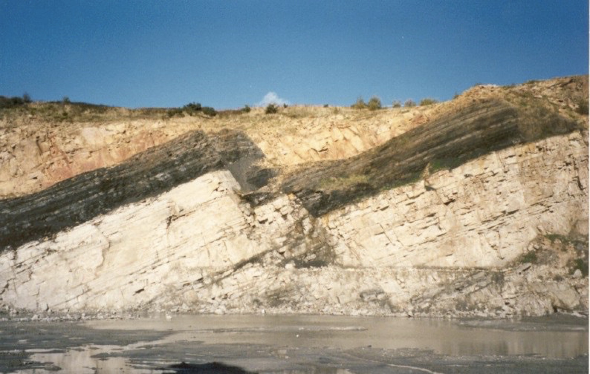 Wall of a quarry with three rock units - white at the base, black in the middle, yellow at the top - that tilt to the right. These formations have all been displaced downwards across two faults in the centre of the picture.