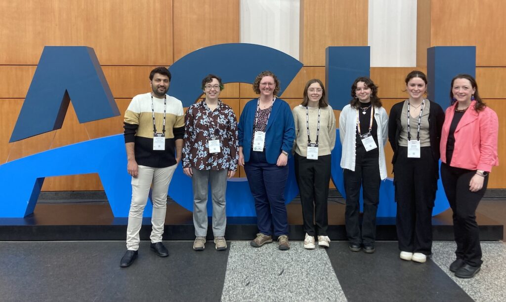 7 people (6 white women and 1 south asian man) stand in front of larger-than-them letters AGU in front of a wooden wall